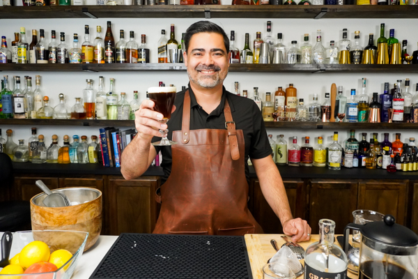 Bartender holding a coffee mug of Jameson Irish Whiskey, 6 ounces hot coffee, and simple syrup for an alcohol-infused coffee drink; on the bar is a coffee pot