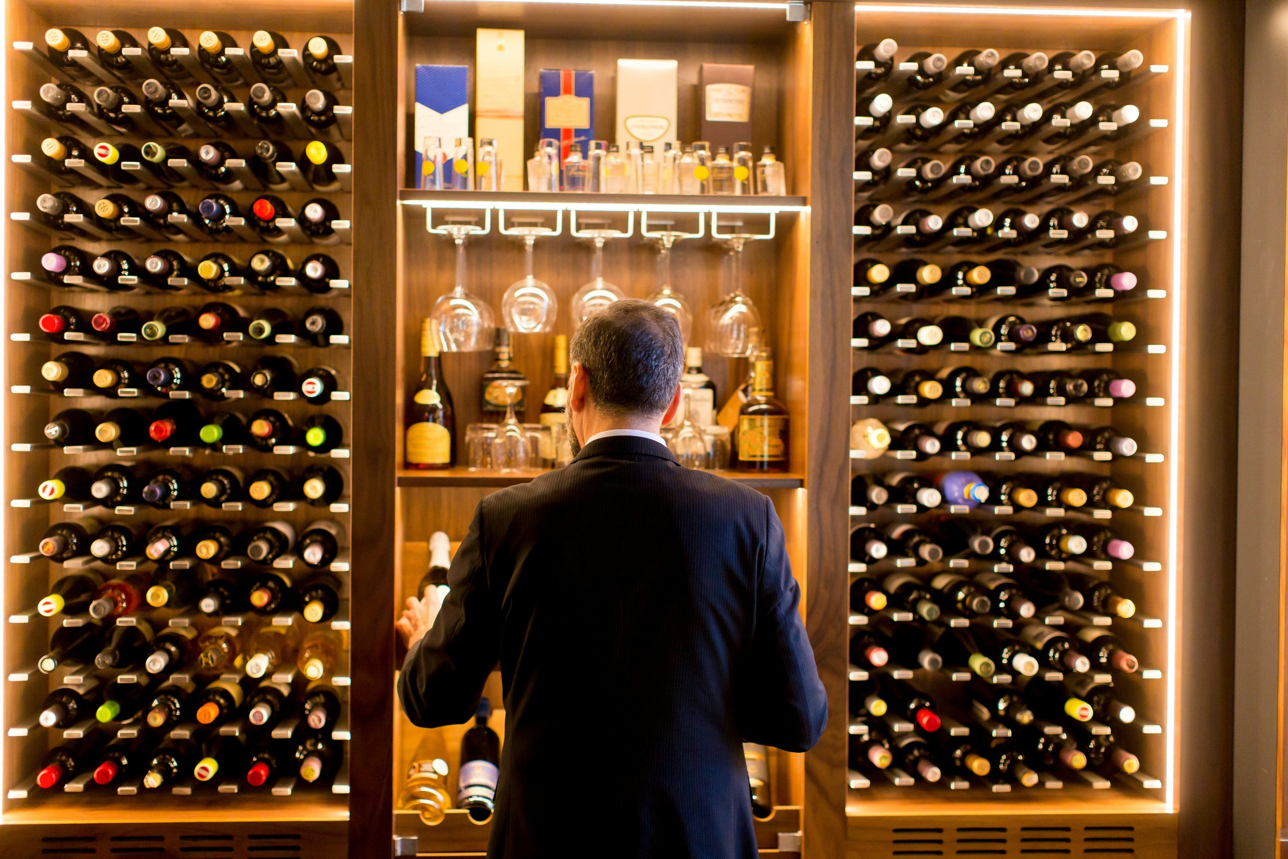Male server in a suit looking at a restaurant wine cellar, photographed from behind