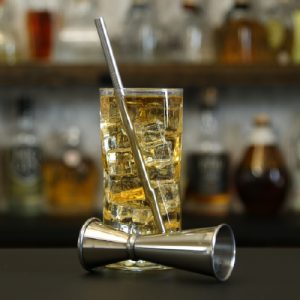 Whiskey highball cocktail in a tall glass with a drinking straw and cocktail jigger