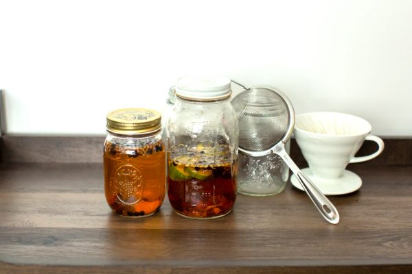 clean Mason jars with liquid that has turned a deep amber color from the aromatic botanicals, next to a fine mesh strainer