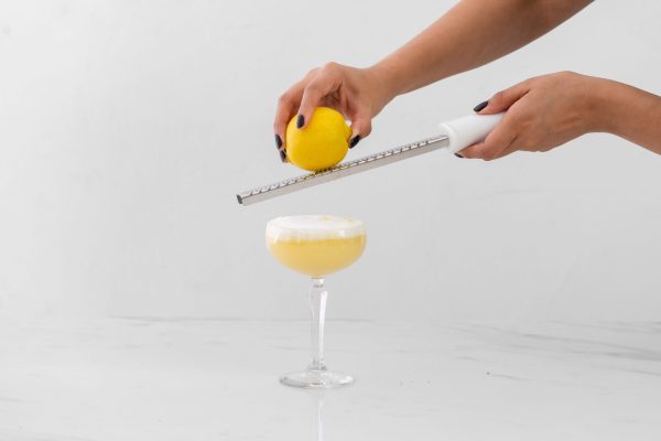 hand holding a kitchen zester with white handle, zesting a lemon over a cocktail